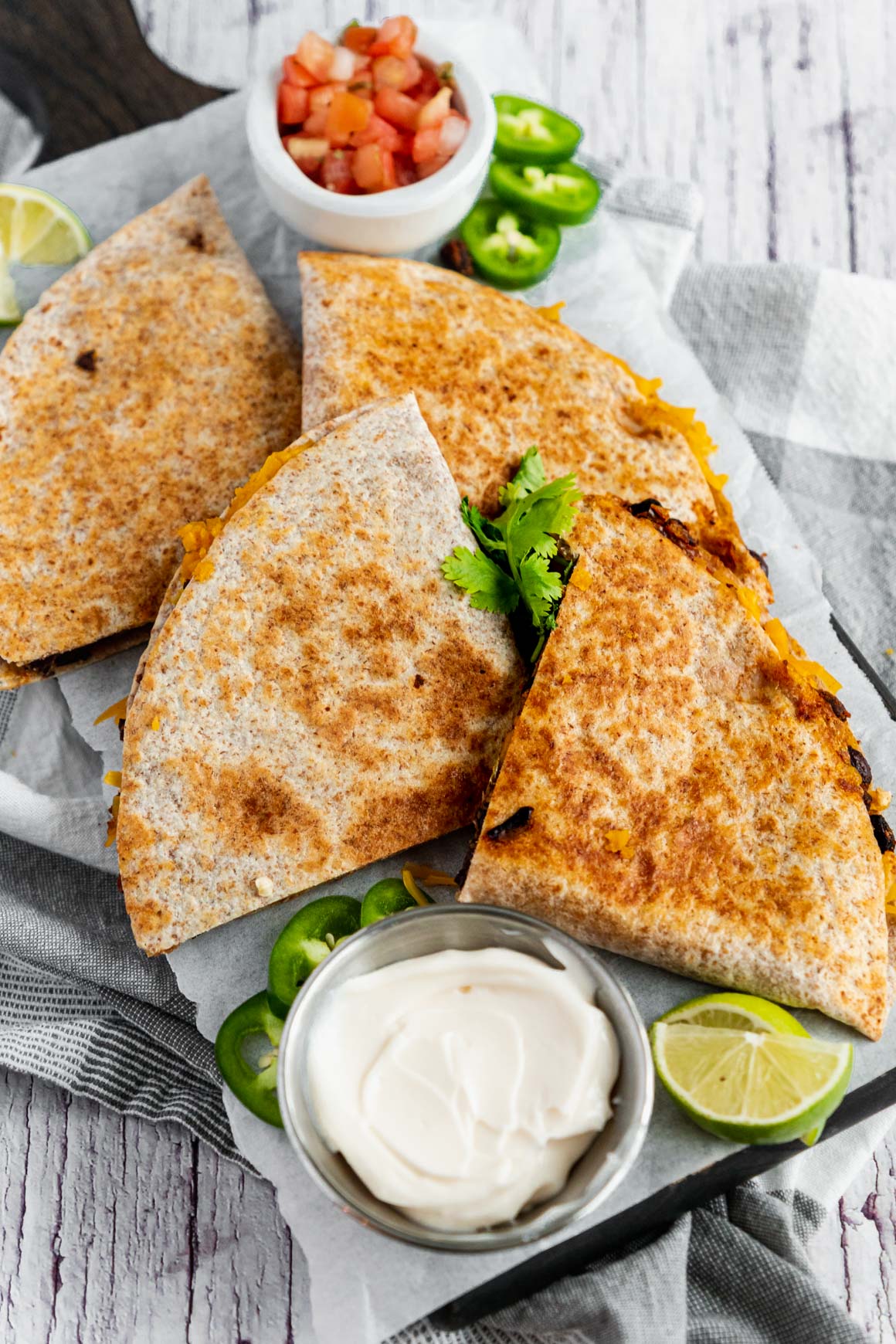 Side-view image of a golden brown black bean quesadilla cute into quarters on a white board on a grey kitchen napkin. To the side there is a small bowl of diced tomato, sliced jalapeno and another bowl of vegan sour cream.