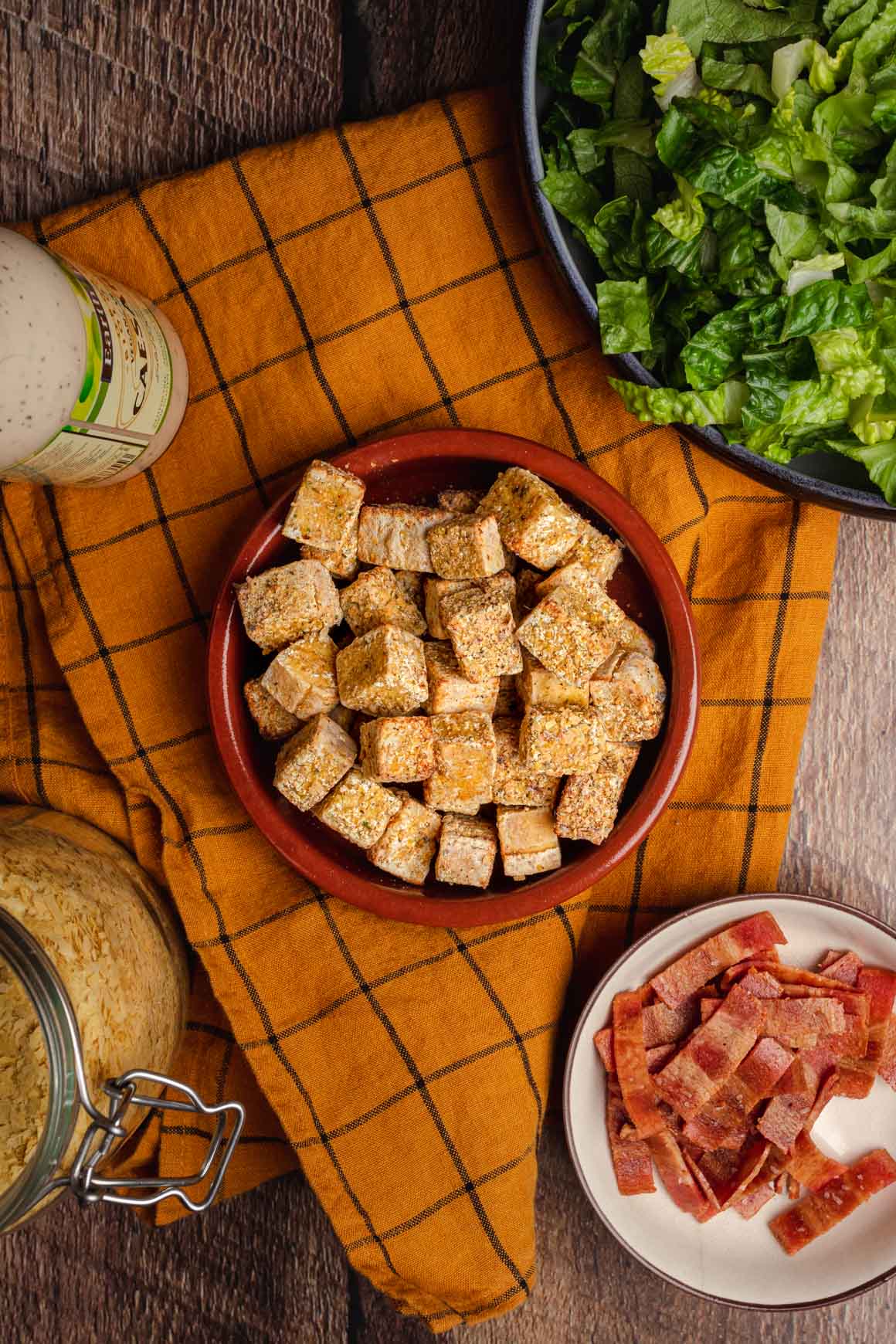 Top down shot of a bowl fo crispy tofu cubes with a yellow cloth underneath. In the corners of the image you can see a bowl of shredded iceberg lettuce, a bowl of crispy vegan bacon, a jar of nutritional yeast, and a bottle of vegan caesar dressing.