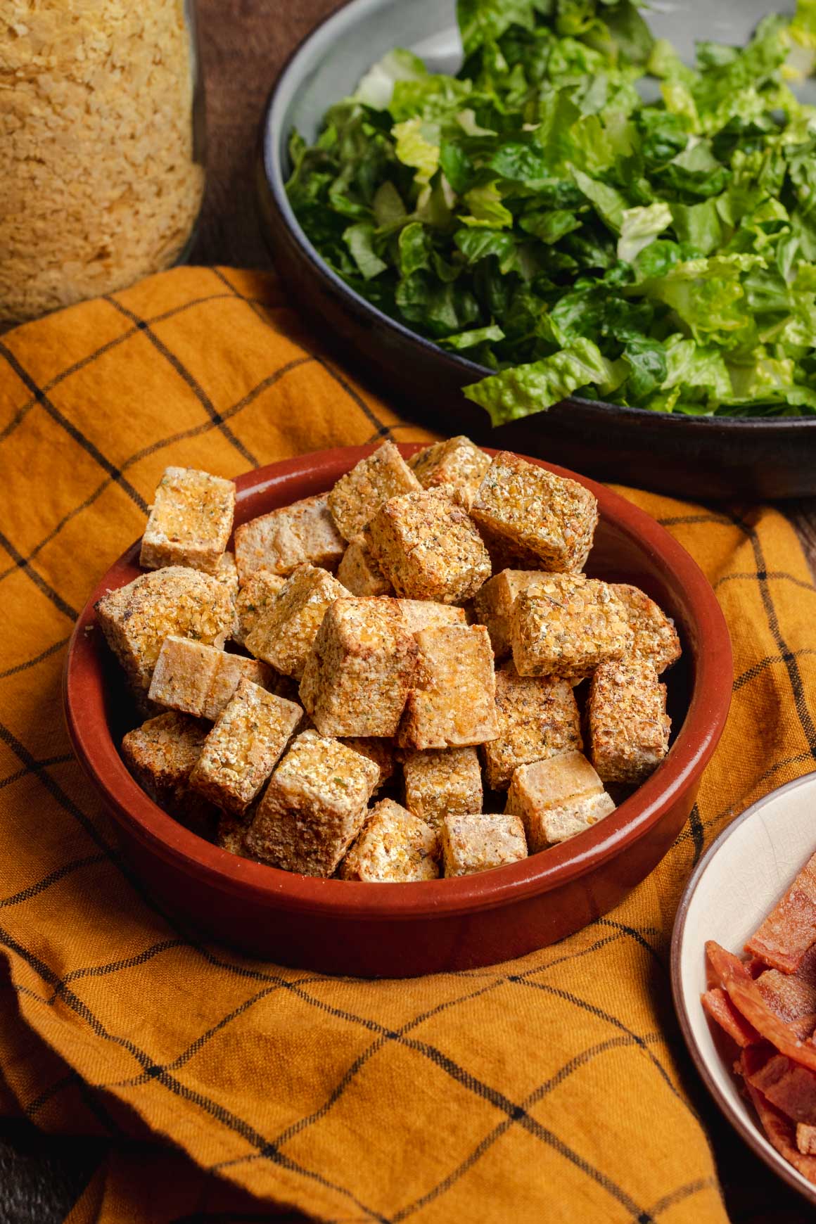 Side view shot of a bowl of crispy tofu cubes with a yellow cloth underneath. In the corners of the image you can see a bowl of shredded iceberg lettuce, a bowl of crispy vegan bacon, and a jar of nutritional yeast.