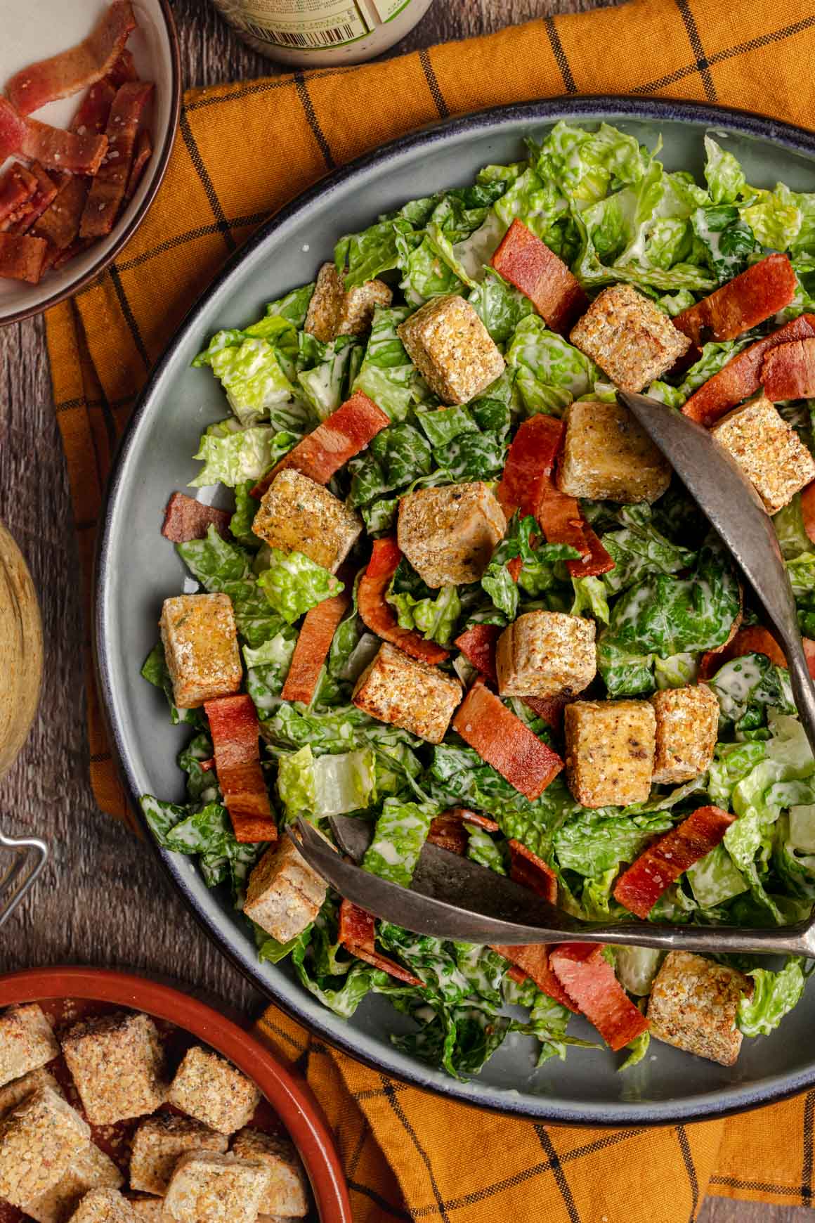 Top down shot of a shallow blue bowl with vegan caesar salad with vegan bacon strips and herby crispy tofu croutons on top of a yellow cloth.