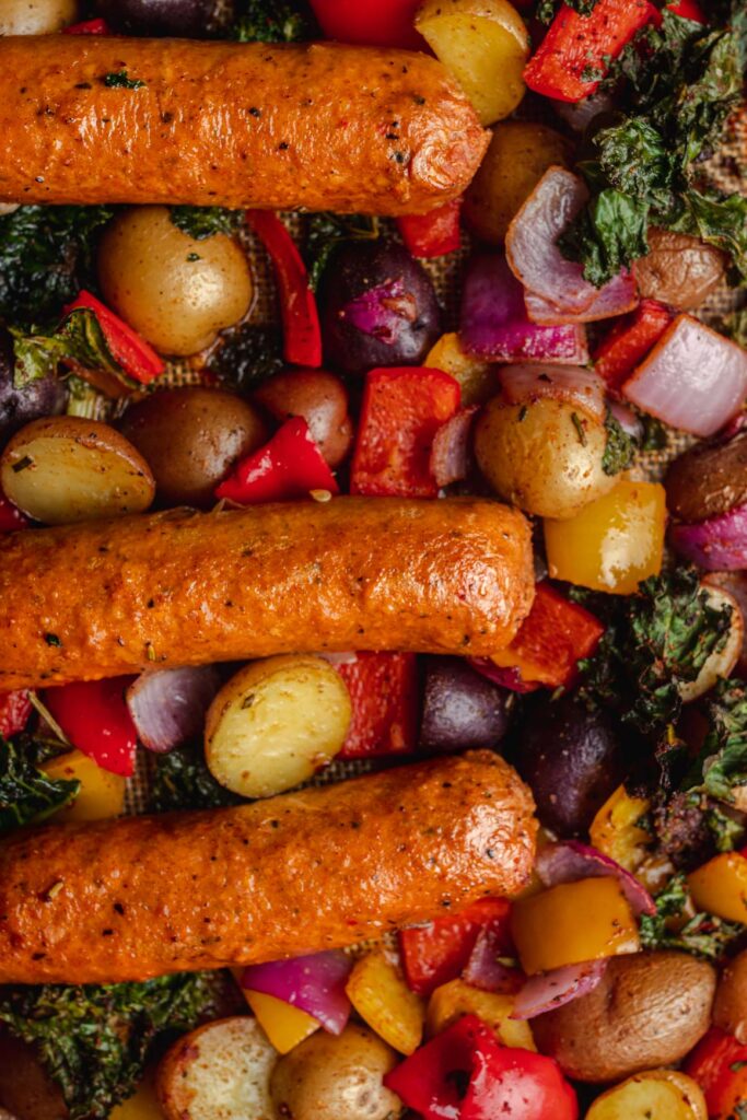 A dark sheet pan peeking in the top of the frame; with roasted bell peppers, baked baby potatoes, crispy kale, and red onion, with beyond sausage on top.