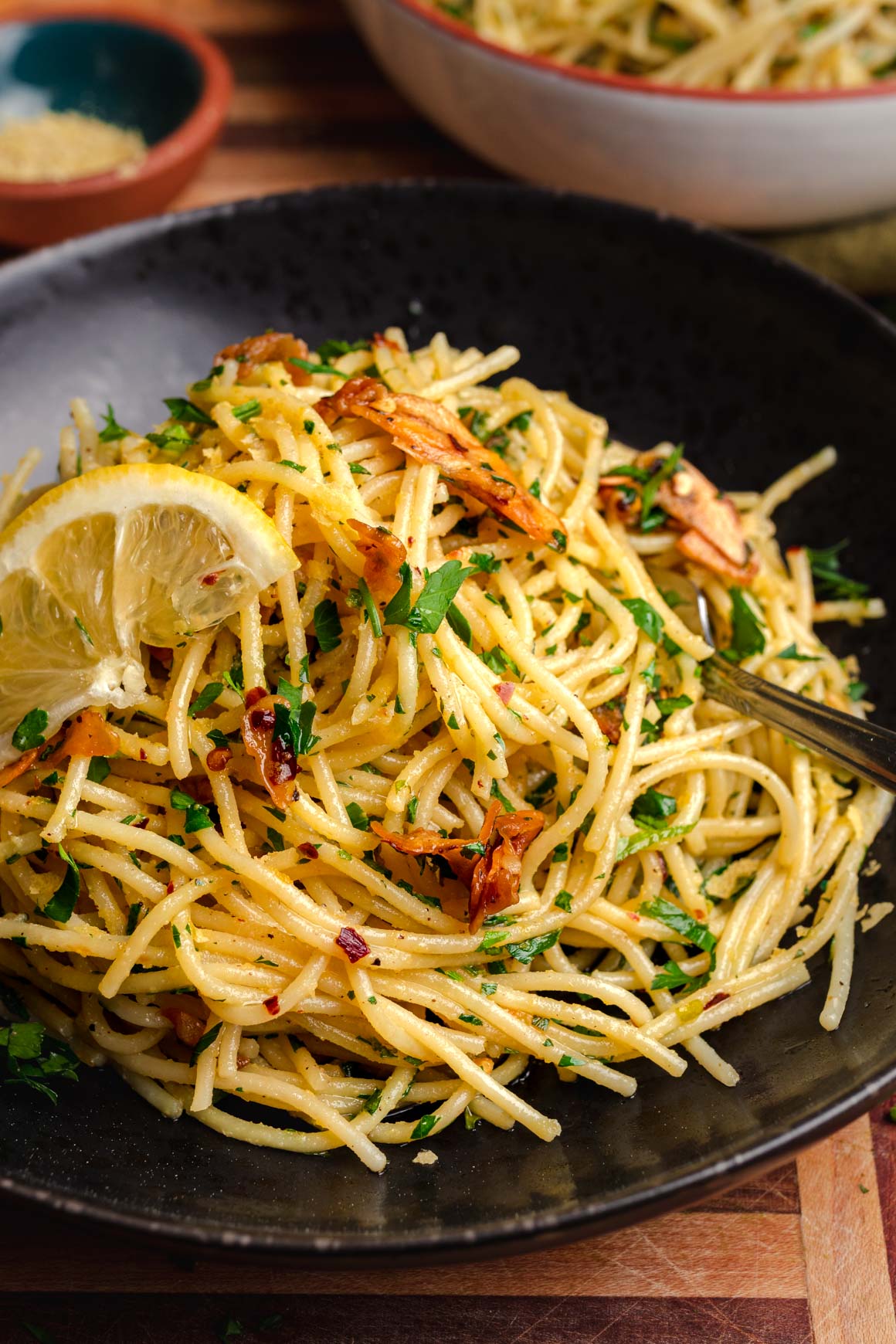 bowl of pasta aglio e olio on a wooden board with minced parsley, garlic cloves and lemon wedges