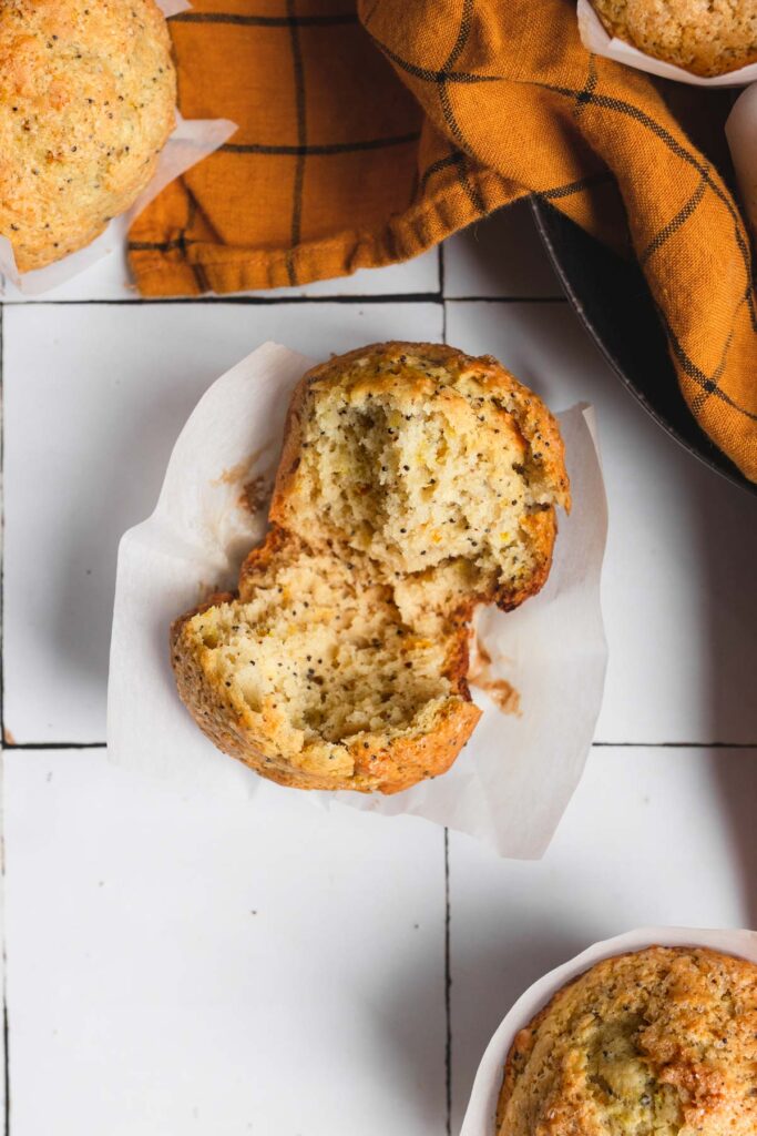 A top-down close up image of a lemon poppy seed muffin split in half on a white tile backdrop. 