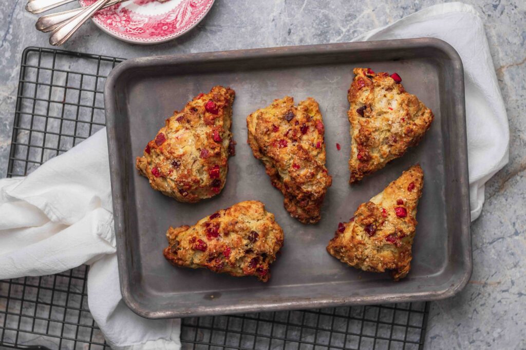 Golden brown fruitcake scones on a small baking sheet on wire rack, with a white linen cloth and a stack of red plates in the upper left corner.