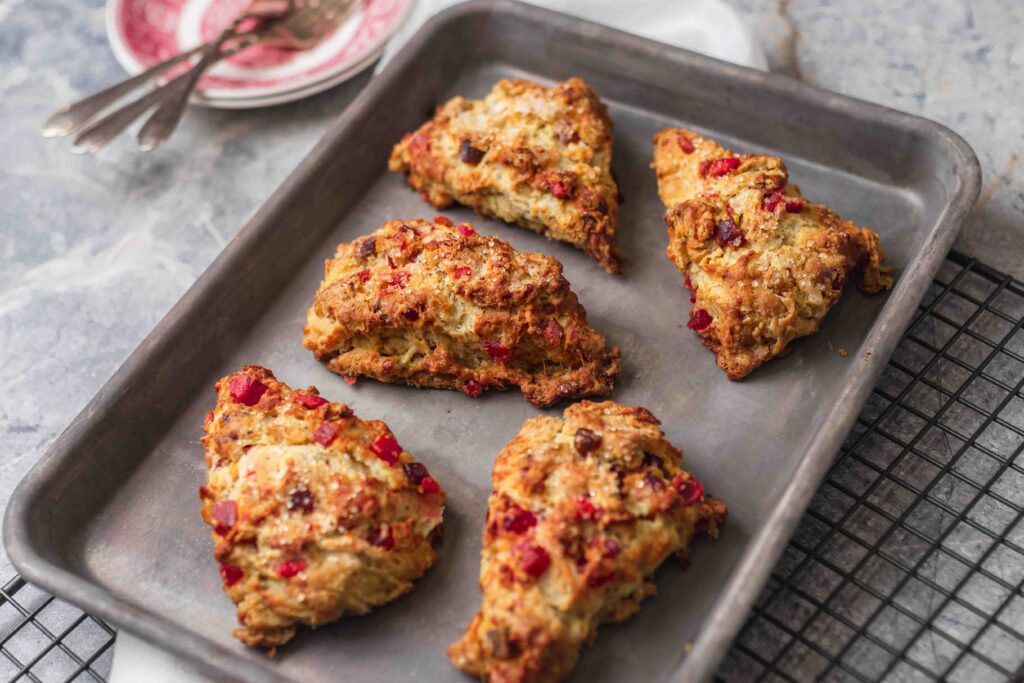 Golden brown fruitcake scones on a small baking sheet on wire rack, with a white linen cloth and a stack of red plates in the upper left corner.