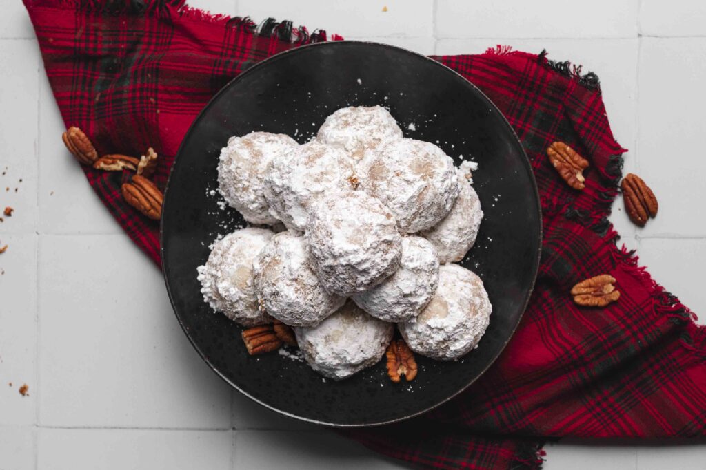 A pile of round pecan puff cookies tossed in icing sugar on a black plate on a red plaid kitchen linen.