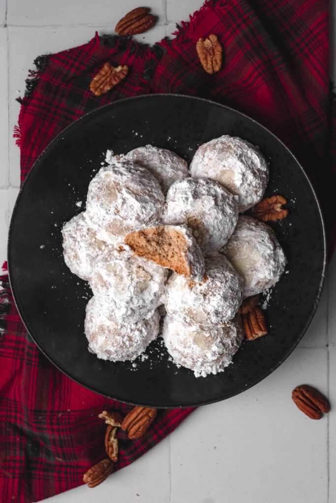A pile of round pecan puff cookies tossed in icing sugar with one on top with a bite out of it on a black plate on a red plaid kitchen linen.