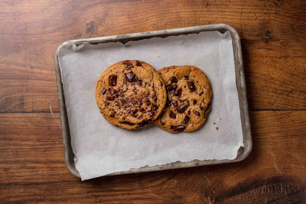 Two large chocolate chunk cookies on a parchment linked aluminum baking sheet on a wood table.
