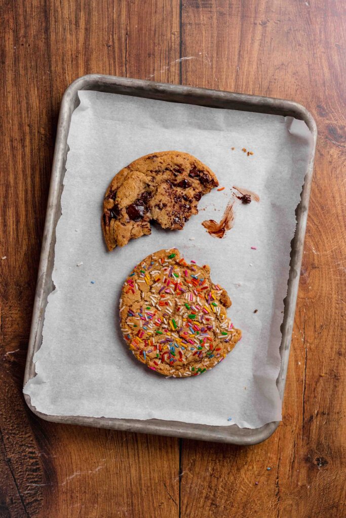 Two half eaten cookies; one chocolate chunk and one sprinkle, on a parchment lined baking sheet on a wood table. 