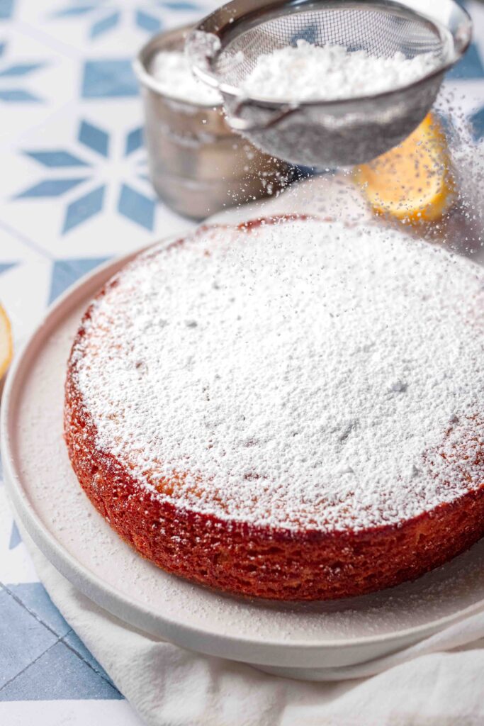 A round vegan lemon cake on a plate with a icing sugar on top and more being sprinkled on top from a sieve. 