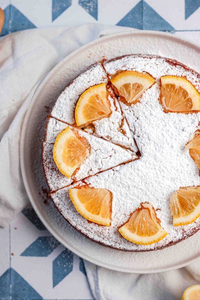Looking down on a round cake on a white plate. The cake is decorated with icing sugar and slices of lemon in a circle around the edge. The cake has two slices cut into it. 