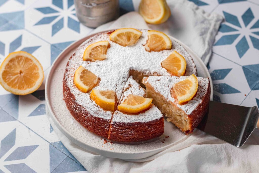Wide image of round lemon cake with icing sugar and lemon slices on top. Two slices cut into the cake with one being lifted by a cake server. 